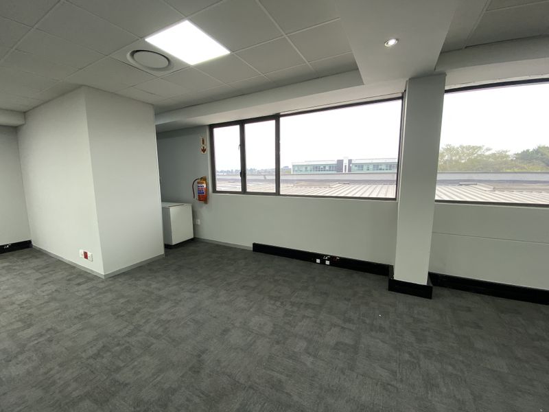 A bright, open-plan office space featuring carpet tiles, suspended ceilings with recessed lighting, and large windows providing natural light.