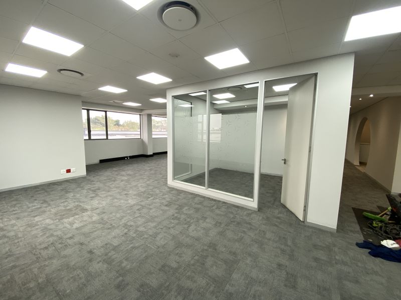 A bright, open-plan office space featuring carpet tiles, a glass-partitioned meeting room, and modern recessed lighting throughout.