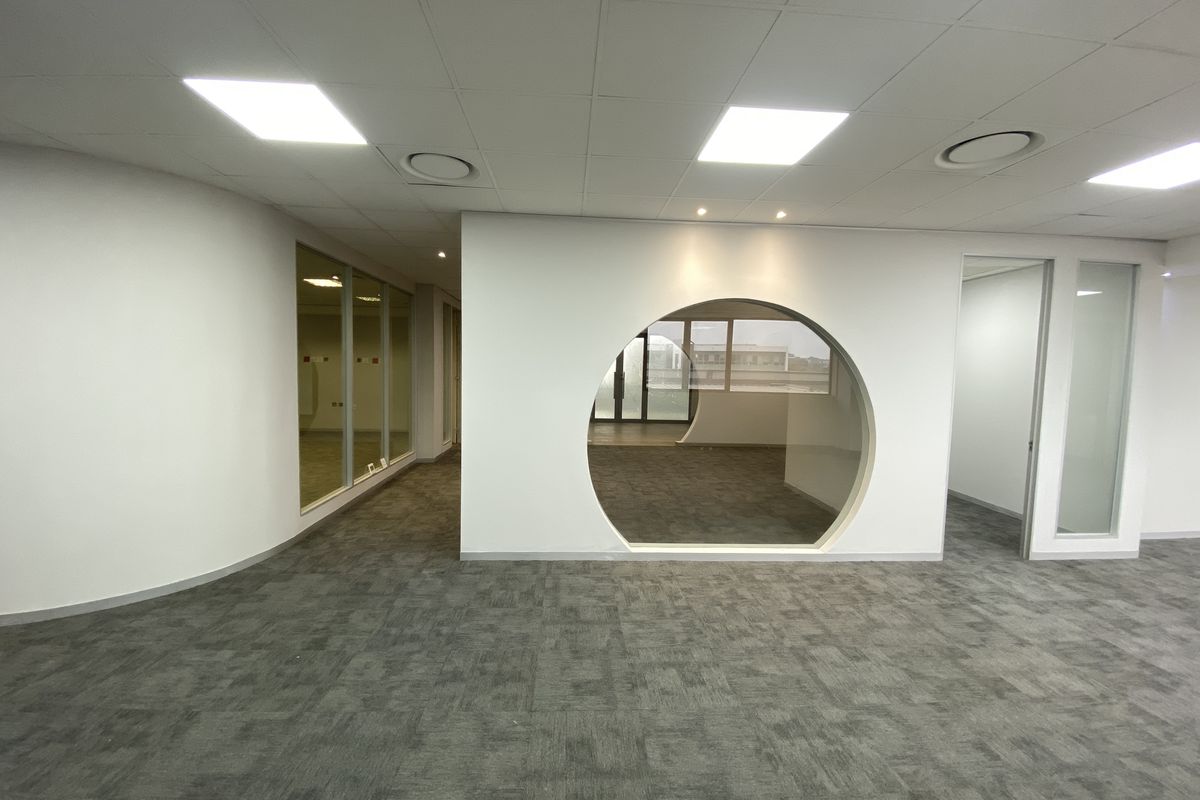 A modern, open-plan office space featuring carpet tiles, a distinctive circular feature window, glass partitions, and recessed ceiling lighting.