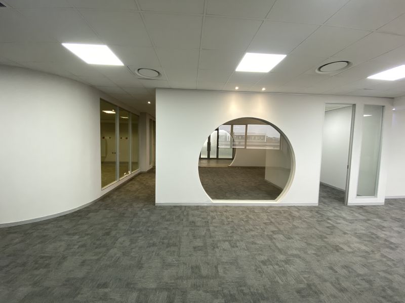 A modern, open-plan office space featuring carpet tiles, a distinctive circular feature window, glass partitions, and recessed ceiling lighting.