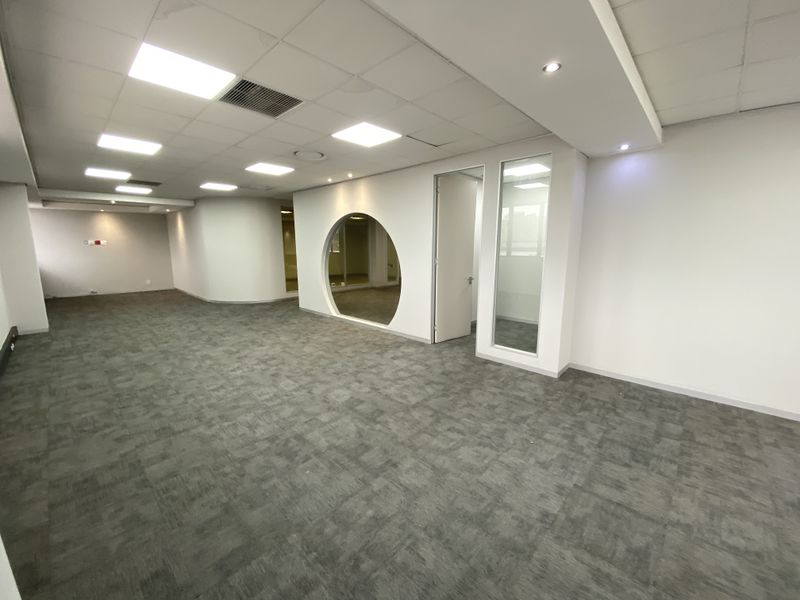 A spacious, empty office area featuring modern carpet tiles, a suspended ceiling with recessed lighting, and a unique circular feature window.