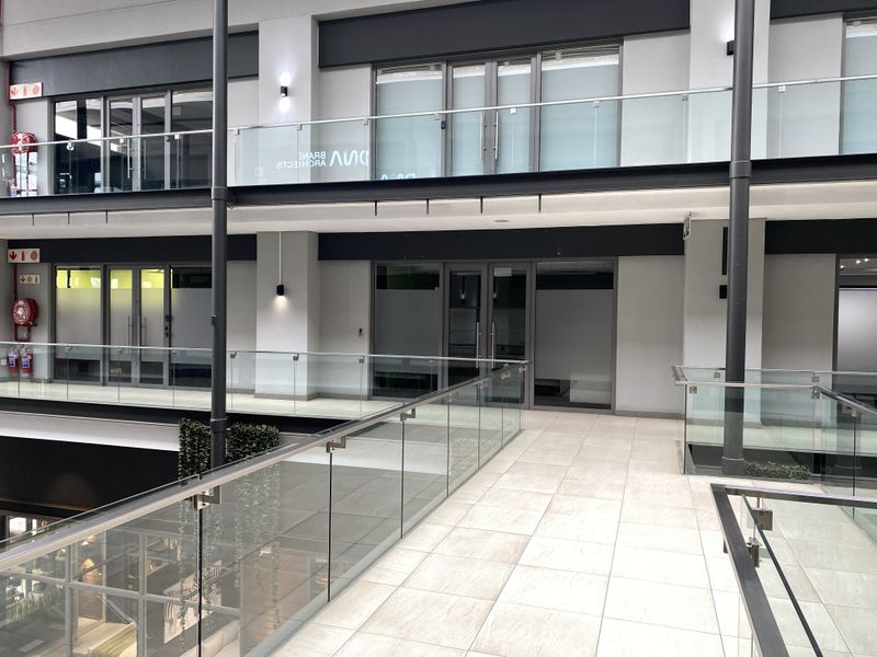 Modern office building interior hallway featuring tiled flooring, glass balustrades, and multiple glass-fronted office entrances across two levels.