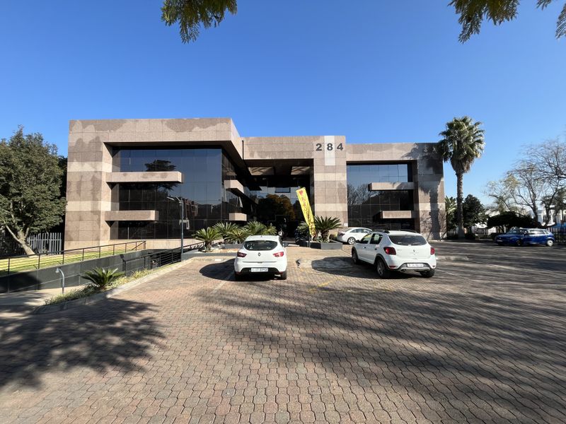 Front elevation of a modern multi-story office building featuring a stone-clad facade, extensive glass curtain walling, and a paved parking area.