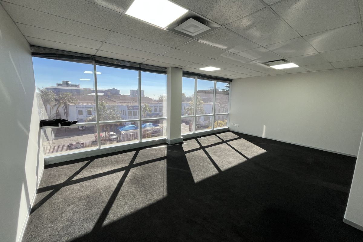 A bright, empty office space featuring large floor-to-ceiling windows, a suspended ceiling with recessed LED lighting, and dark carpeted flooring.