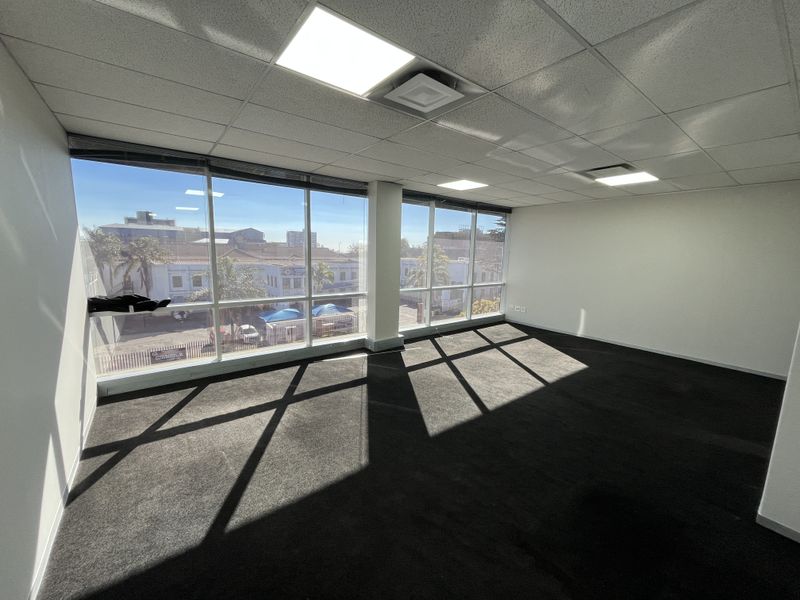 A bright, empty office space featuring large floor-to-ceiling windows, a suspended ceiling with recessed LED lighting, and dark carpeted flooring.
