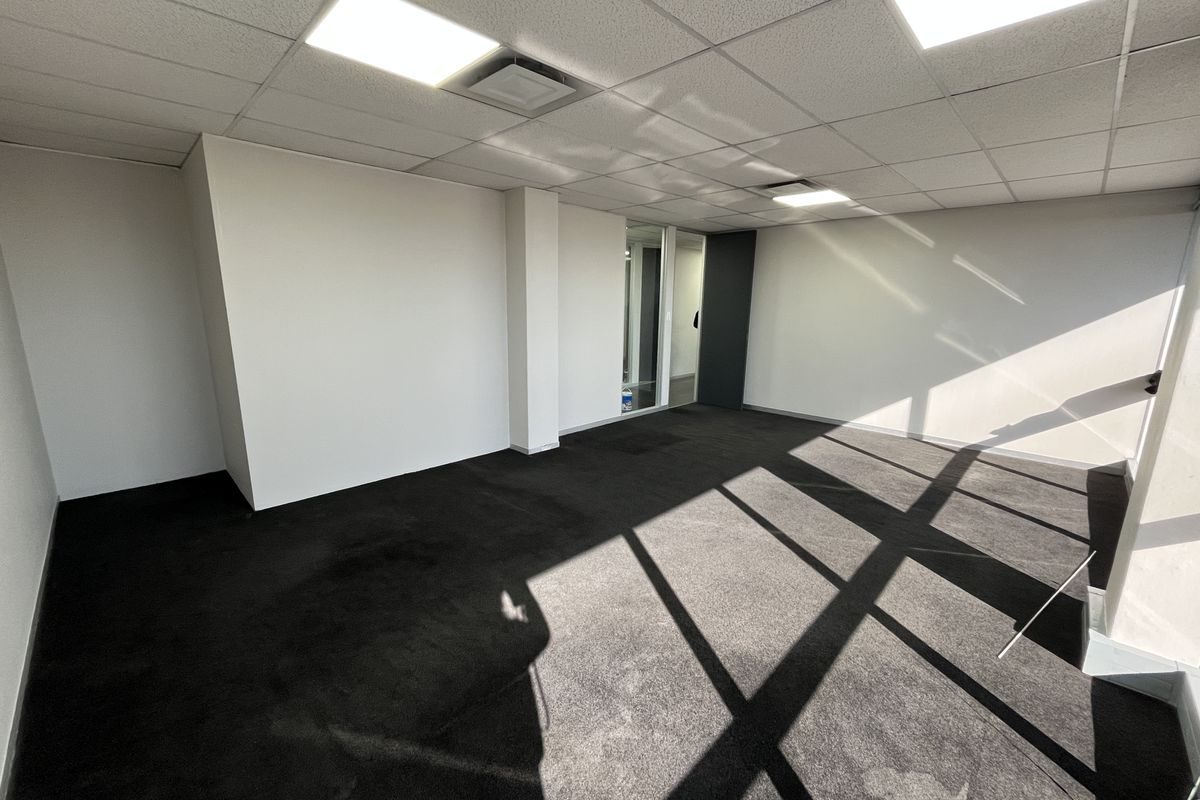 A vacant, open-plan office space featuring a suspended ceiling with recessed lighting, dark carpeting, and a glass partition wall.