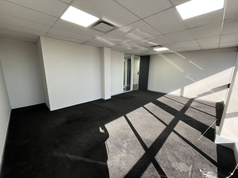 A vacant, open-plan office space featuring a suspended ceiling with recessed lighting, dark carpeting, and a glass partition wall.