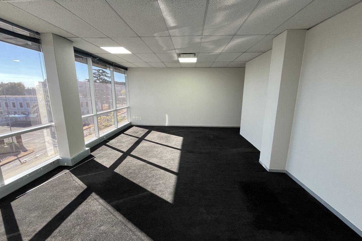 A bright, empty office space featuring large windows, a suspended ceiling with recessed lighting, and dark carpeted flooring.
