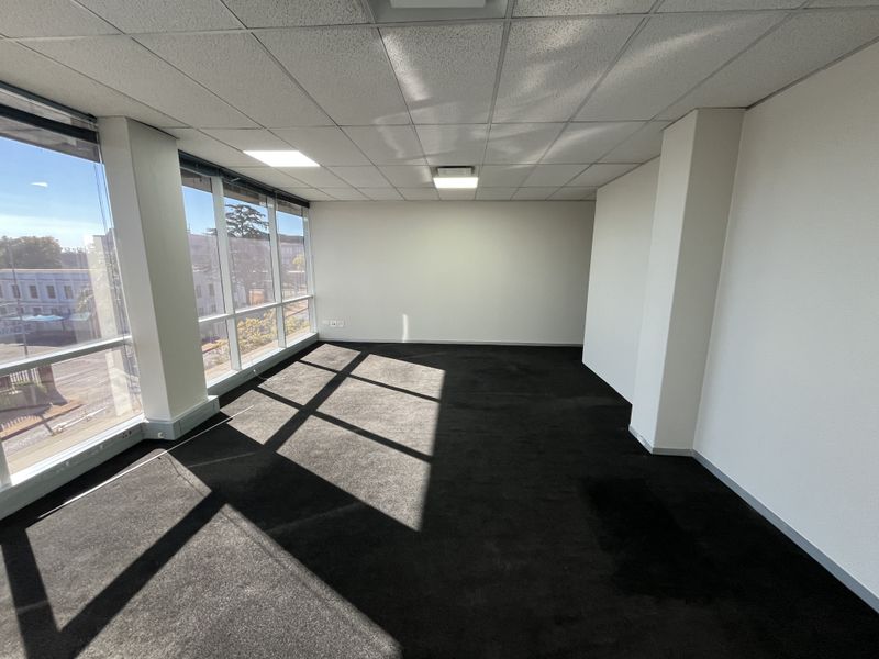 A bright, empty office space featuring large windows, a suspended ceiling with recessed lighting, and dark carpeted flooring.