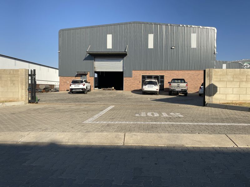 Front elevation of an industrial warehouse featuring a large roller shutter door, brick and metal cladding facade, and a paved driveway with secure gated access.