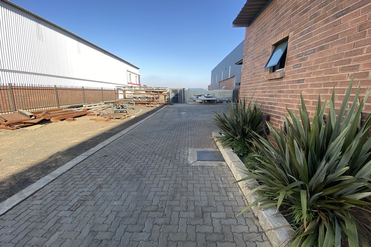 A paved industrial rear yard area featuring brick and metal building facades, security fencing, and established landscaping.