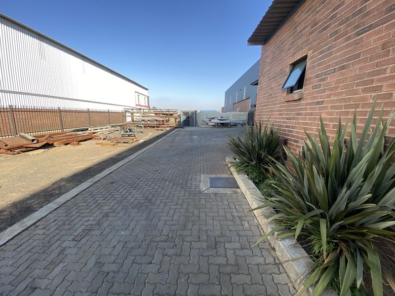 A paved industrial rear yard area featuring brick and metal building facades, security fencing, and established landscaping.