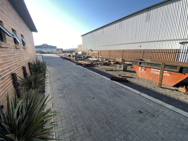 Exterior view of an industrial property featuring a paved driveway, brick building facade, and corrugated metal warehouse structure with adjacent yard space.