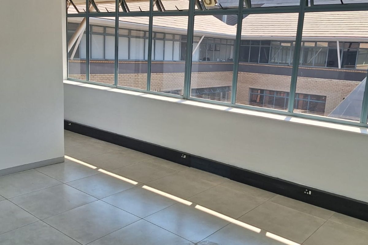 A bright, empty office space featuring large windows, tiled flooring, and integrated electrical trunking along the base of the wall.