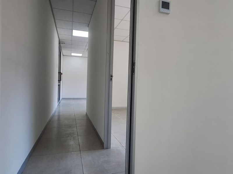 A clean, empty office hallway featuring tiled flooring, a suspended ceiling with recessed lighting, and a wall-mounted thermostat.