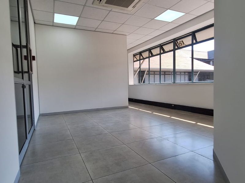 A bright, empty office space featuring tiled flooring, a suspended ceiling with recessed lighting, and large windows providing natural light.