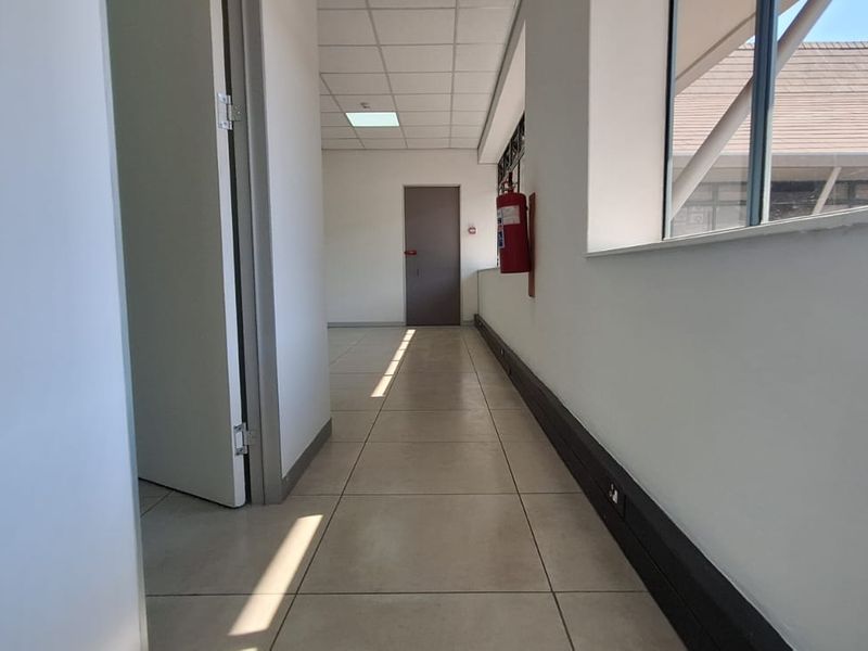 A bright, empty office hallway featuring tiled flooring, a suspended ceiling, and large windows providing natural light.
