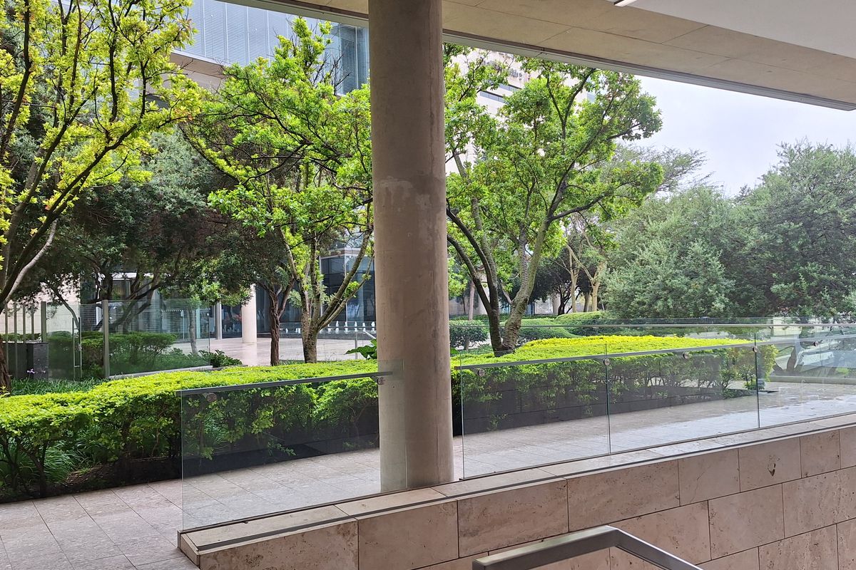 A modern covered outdoor terrace featuring stone tile flooring, a glass balustrade, and clean architectural lines overlooking a landscaped garden area.