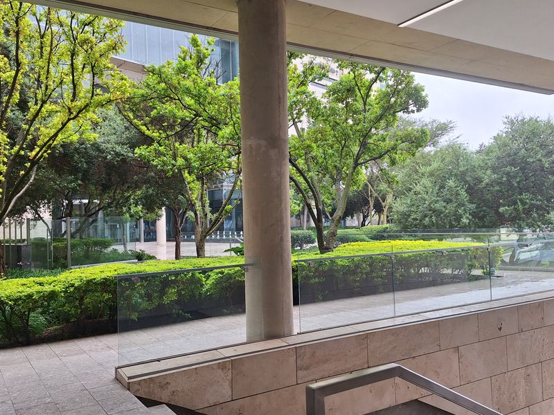A modern covered outdoor terrace featuring stone tile flooring, a glass balustrade, and clean architectural lines overlooking a landscaped garden area.