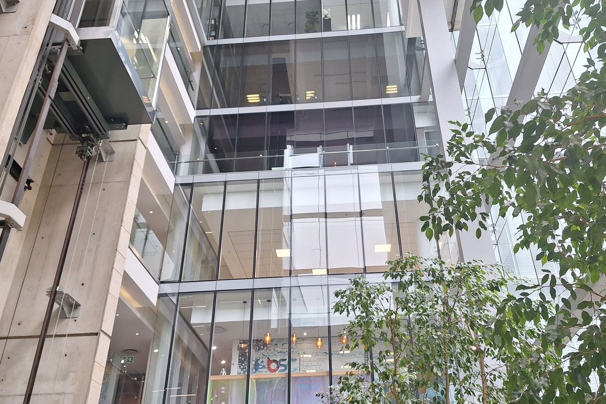 Modern multi-story office atrium featuring a glass curtain wall, exposed concrete structural elements, and a prominent glass elevator shaft.