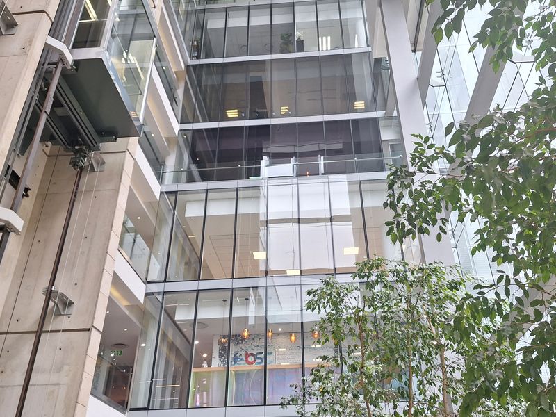 Modern multi-story office atrium featuring a glass curtain wall, exposed concrete structural elements, and a prominent glass elevator shaft.