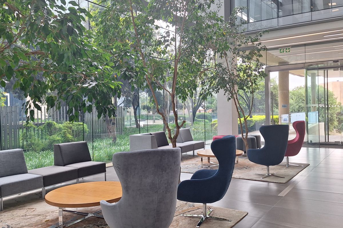 Modern office building lobby featuring floor-to-ceiling glass walls, tiled flooring, and a bright, open-plan reception area with integrated indoor greenery.
