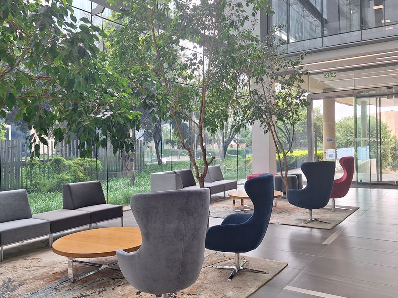 Modern office building lobby featuring floor-to-ceiling glass walls, tiled flooring, and a bright, open-plan reception area with integrated indoor greenery.
