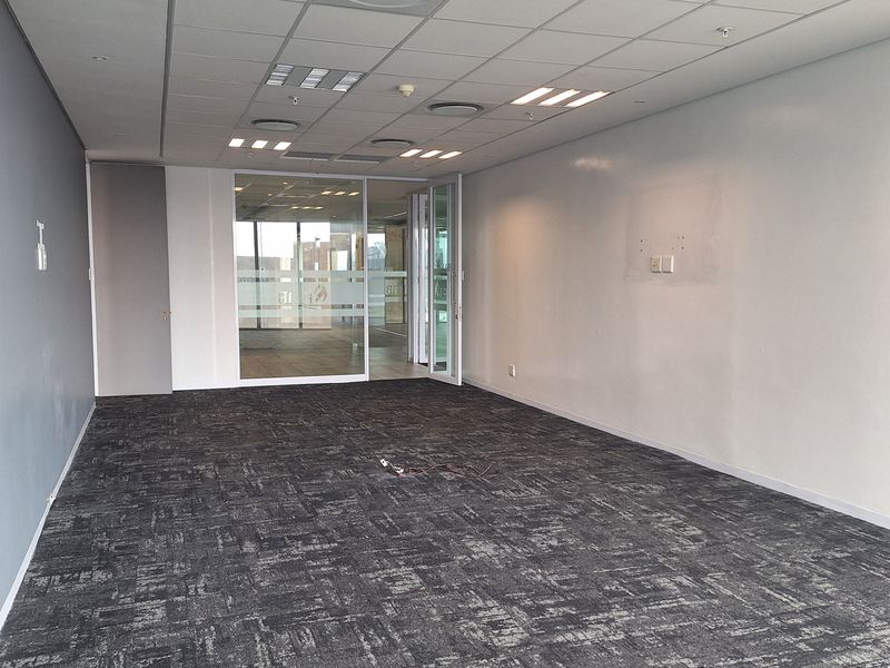 An empty, open-plan office space featuring grey carpet tiles, a suspended ceiling with recessed lighting, and a glass partition leading to an adjacent area.