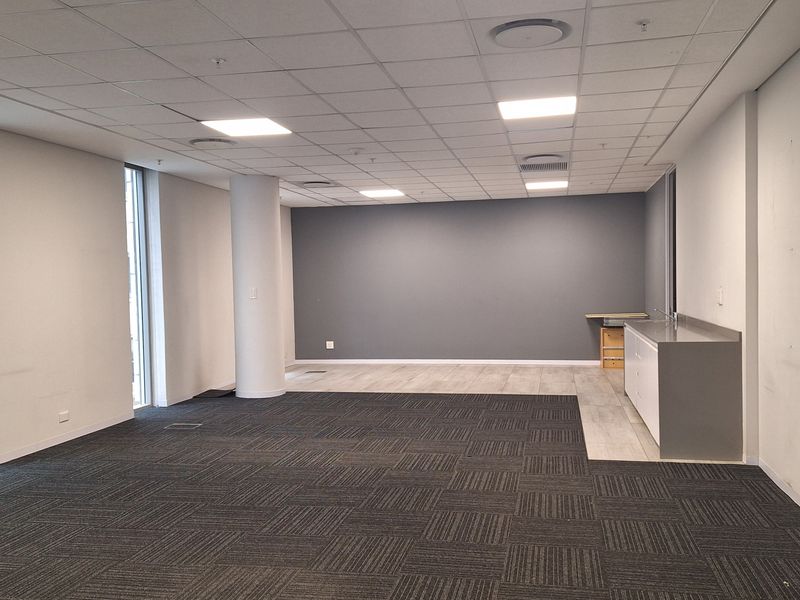 A spacious, modern open-plan office area featuring a suspended ceiling, recessed lighting, carpet tiles, and a compact built-in kitchenette unit.