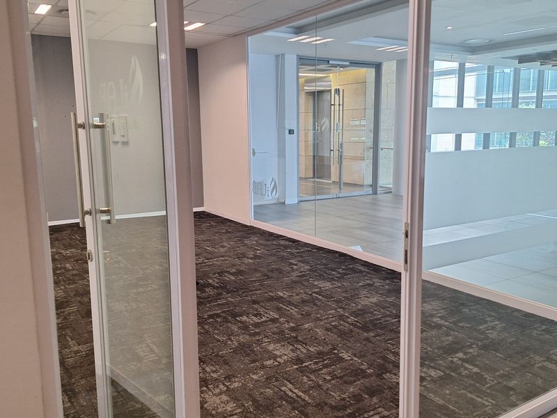 A modern, vacant office space featuring glass partition walls, carpeted flooring, and a suspended ceiling with recessed lighting, offering a professional and bright work environment.