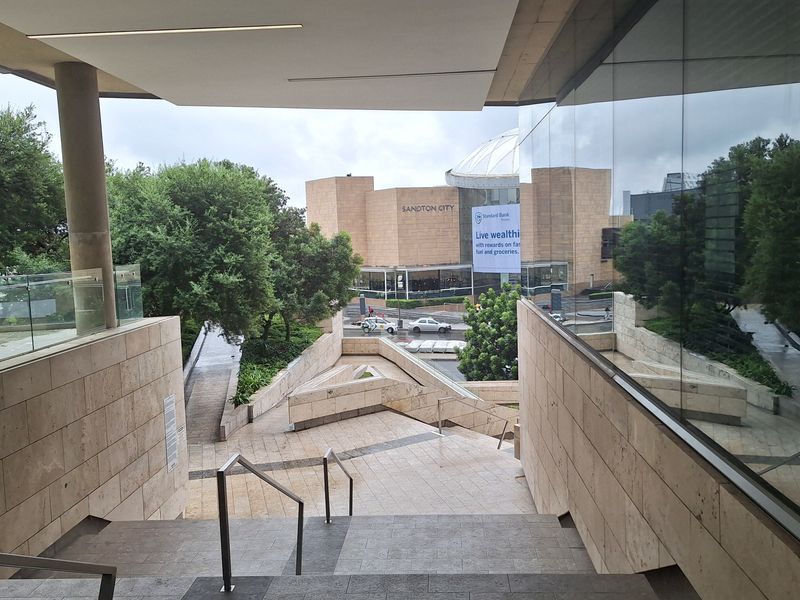 Modern office building entrance featuring stone-clad stairs, glass facade, and architectural metal handrails overlooking a landscaped urban plaza.