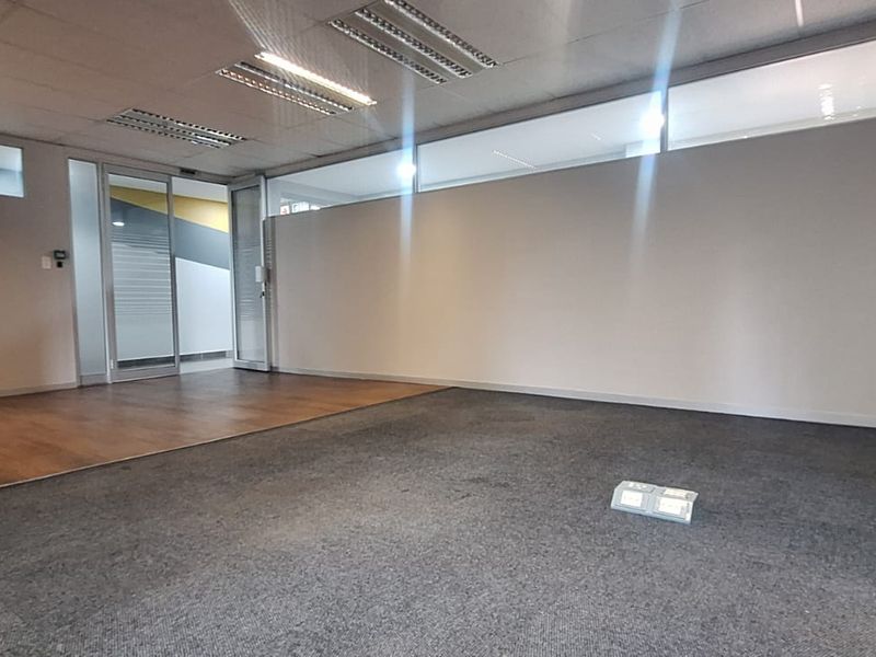 A spacious, empty office area featuring a mix of carpet and laminate flooring, glass partitions, and recessed ceiling lighting.