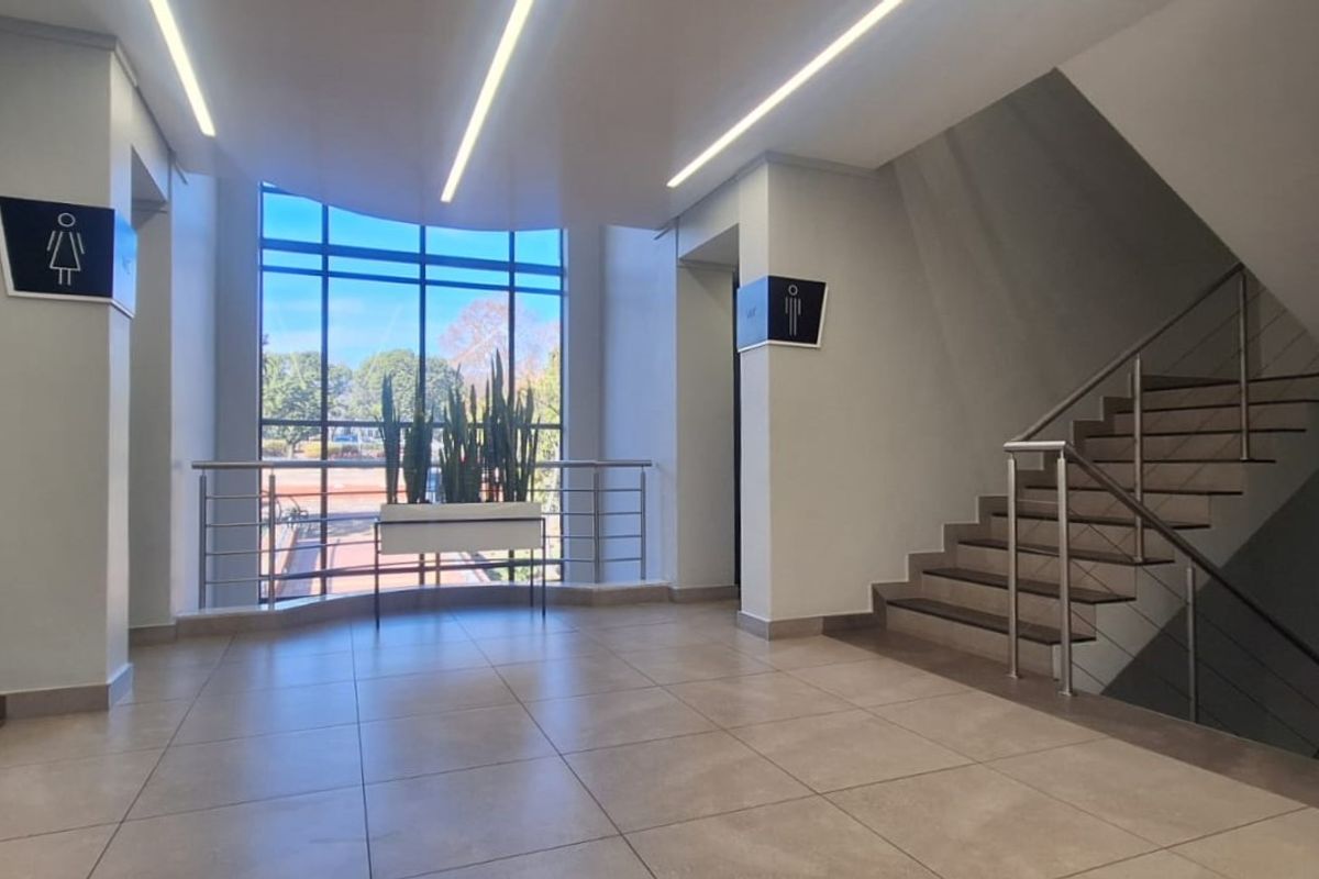 Modern office building hallway featuring tiled flooring, recessed linear lighting, a prominent staircase with metal railings, and large windows providing natural light.