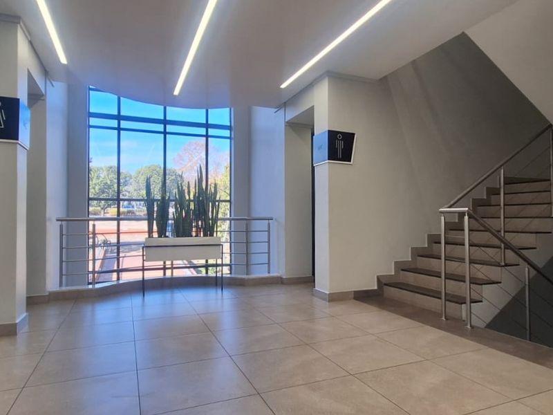 Modern office building hallway featuring tiled flooring, recessed linear lighting, a prominent staircase with metal railings, and large windows providing natural light.
