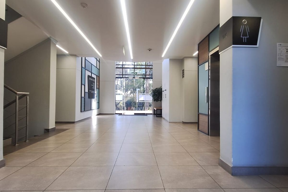 Modern office building lobby featuring tiled flooring, linear LED lighting, elevator access, and a glass entrance door leading to the exterior.