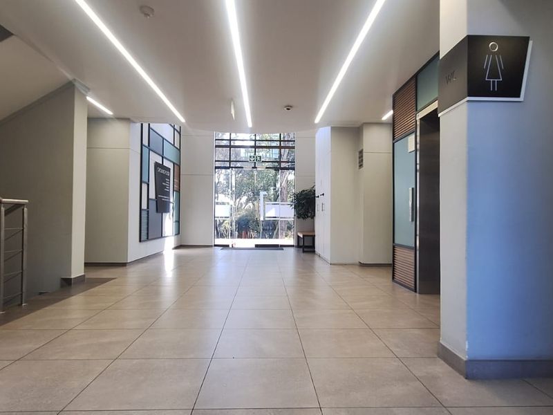 Modern office building lobby featuring tiled flooring, linear LED lighting, elevator access, and a glass entrance door leading to the exterior.