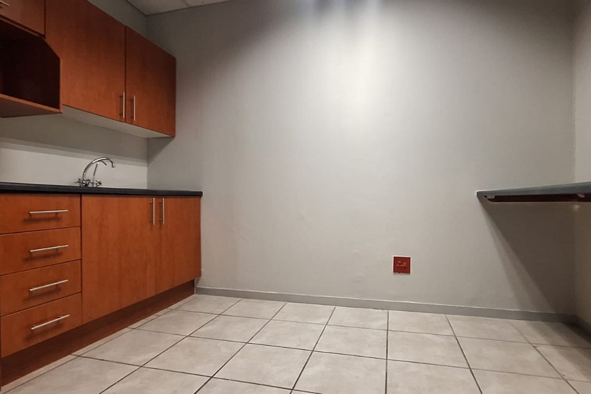 A functional office kitchenette featuring wood-finish cabinetry, a stainless steel sink, laminate countertops, and neutral tiled flooring.