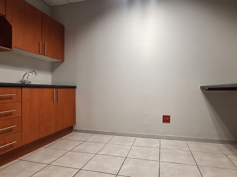 A functional office kitchenette featuring wood-finish cabinetry, a stainless steel sink, laminate countertops, and neutral tiled flooring.