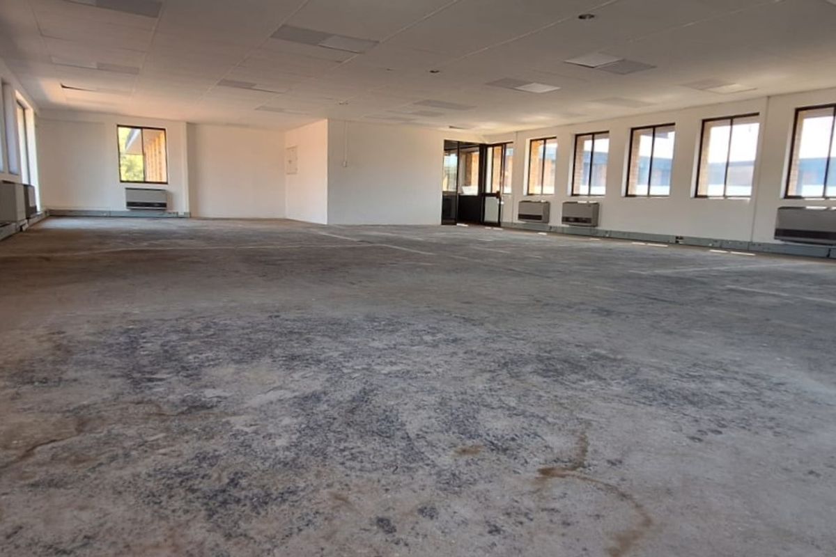 A large, open-plan office space featuring a suspended ceiling, multiple wall-mounted air conditioning units, and large windows providing ample natural light.