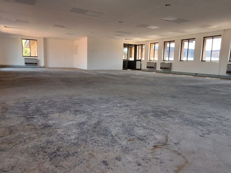 A large, open-plan office space featuring a suspended ceiling, multiple wall-mounted air conditioning units, and large windows providing ample natural light.