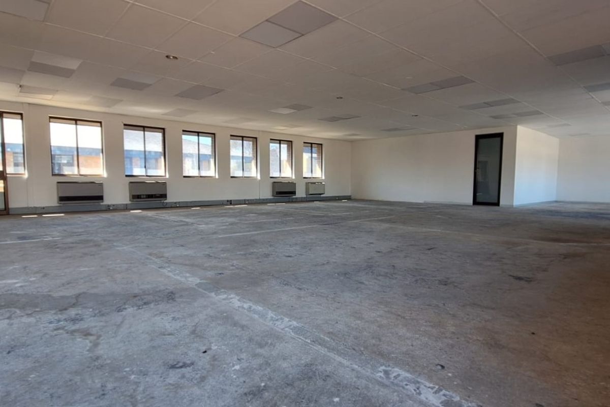 A spacious, vacant open-plan office area featuring large windows, wall-mounted air conditioning units, and a suspended ceiling with exposed concrete flooring.