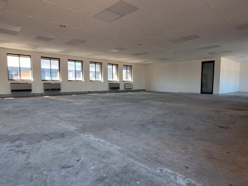 A spacious, vacant open-plan office area featuring large windows, wall-mounted air conditioning units, and a suspended ceiling with exposed concrete flooring.
