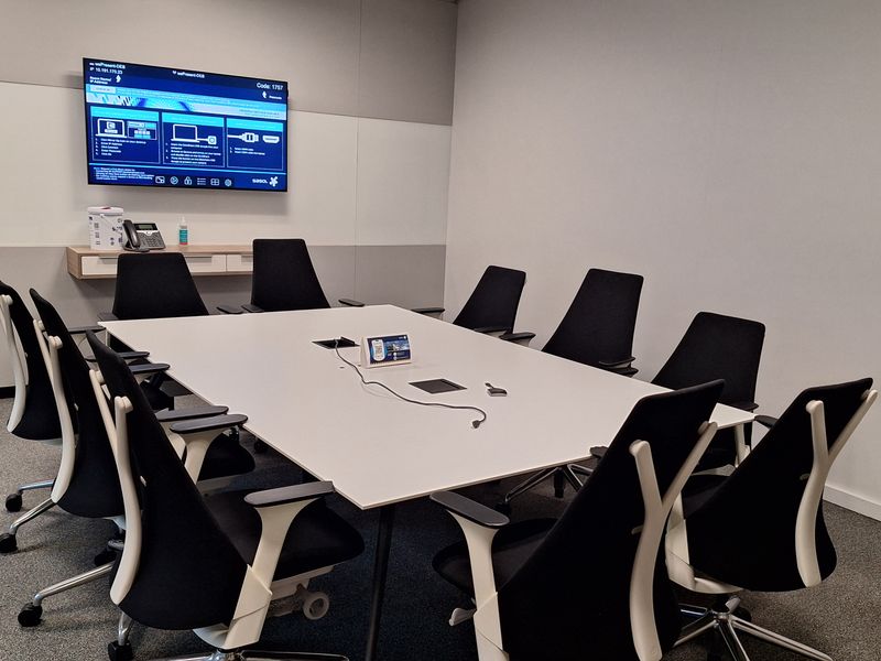 A professional office meeting room featuring a large central conference table, ergonomic seating, and a wall-mounted display screen for presentations.