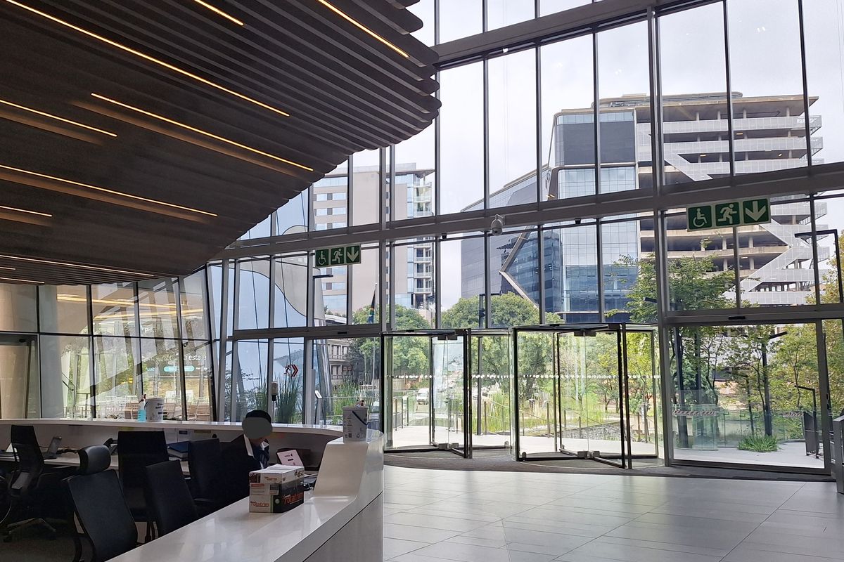 Modern office building entrance lobby featuring a large reception desk, floor-to-ceiling glass facade, and contemporary architectural ceiling design.