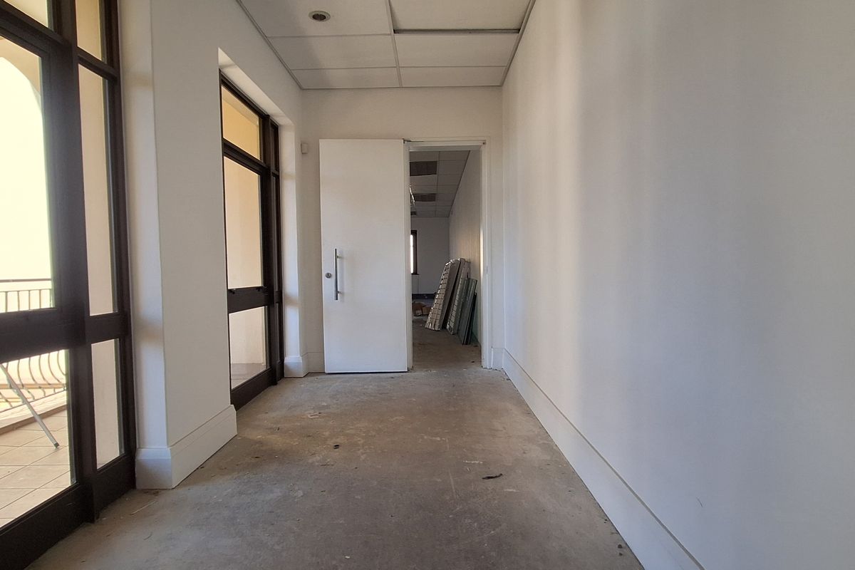 A vacant office hallway featuring a suspended ceiling, recessed lighting, and access to adjacent office spaces through glass-paneled doors.