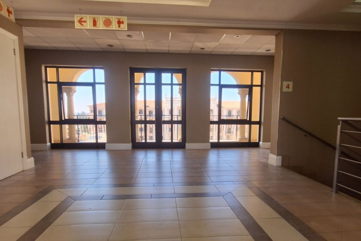 A spacious, well-lit office hallway featuring tiled flooring, a suspended ceiling, and large windows providing natural light and access to an outdoor balcony area.