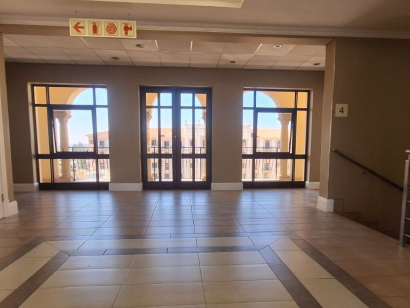 A spacious, well-lit office hallway featuring tiled flooring, a suspended ceiling, and large windows providing natural light and access to an outdoor balcony area.