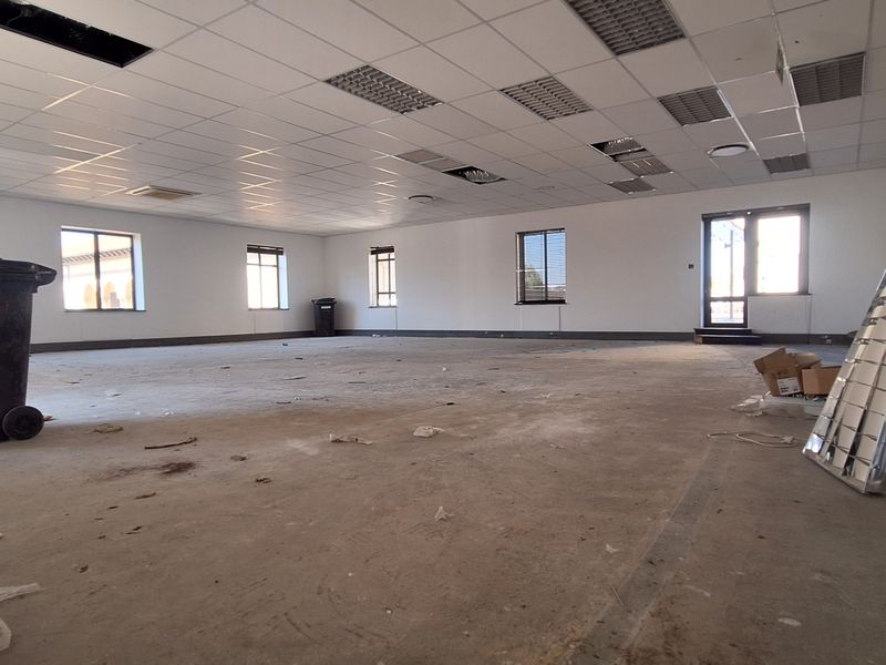 A large, open-plan office space featuring a suspended ceiling with recessed lighting and multiple windows, currently in need of flooring and ceiling repairs.