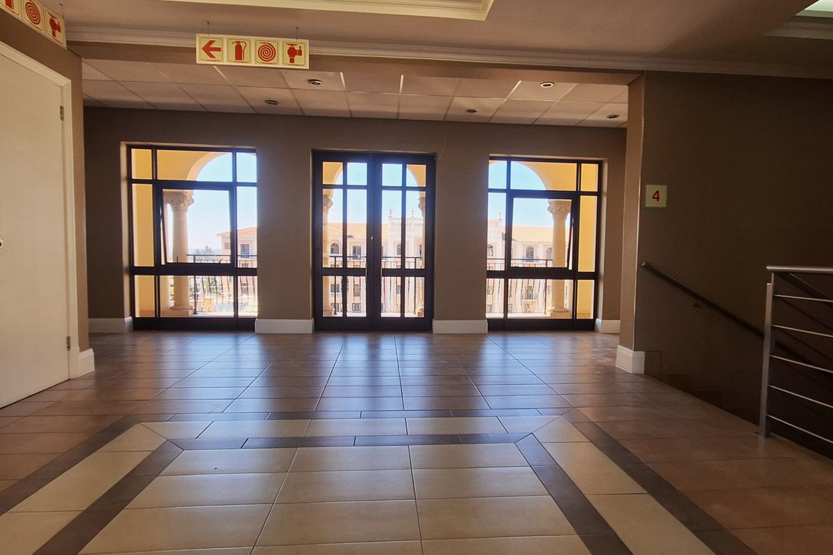 A spacious, well-maintained office building hallway featuring tiled floors, a suspended ceiling, and large windows providing natural light and access to a balcony.