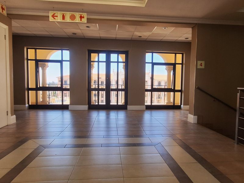 A spacious, well-maintained office building hallway featuring tiled floors, a suspended ceiling, and large windows providing natural light and access to a balcony.